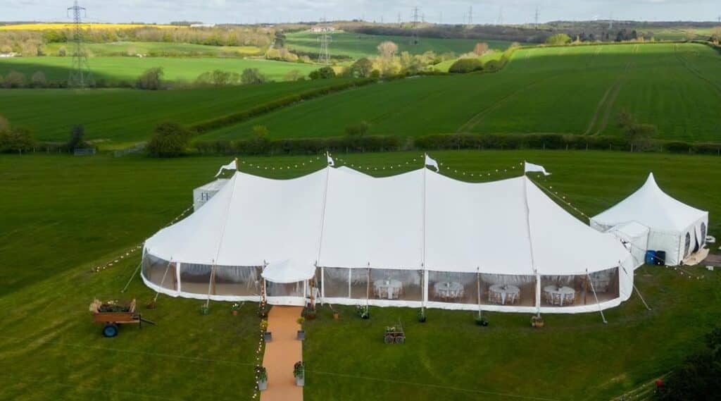 A large white marquee tent on a grassy field with string lights and a smaller adjacent tent in a rural landscape.