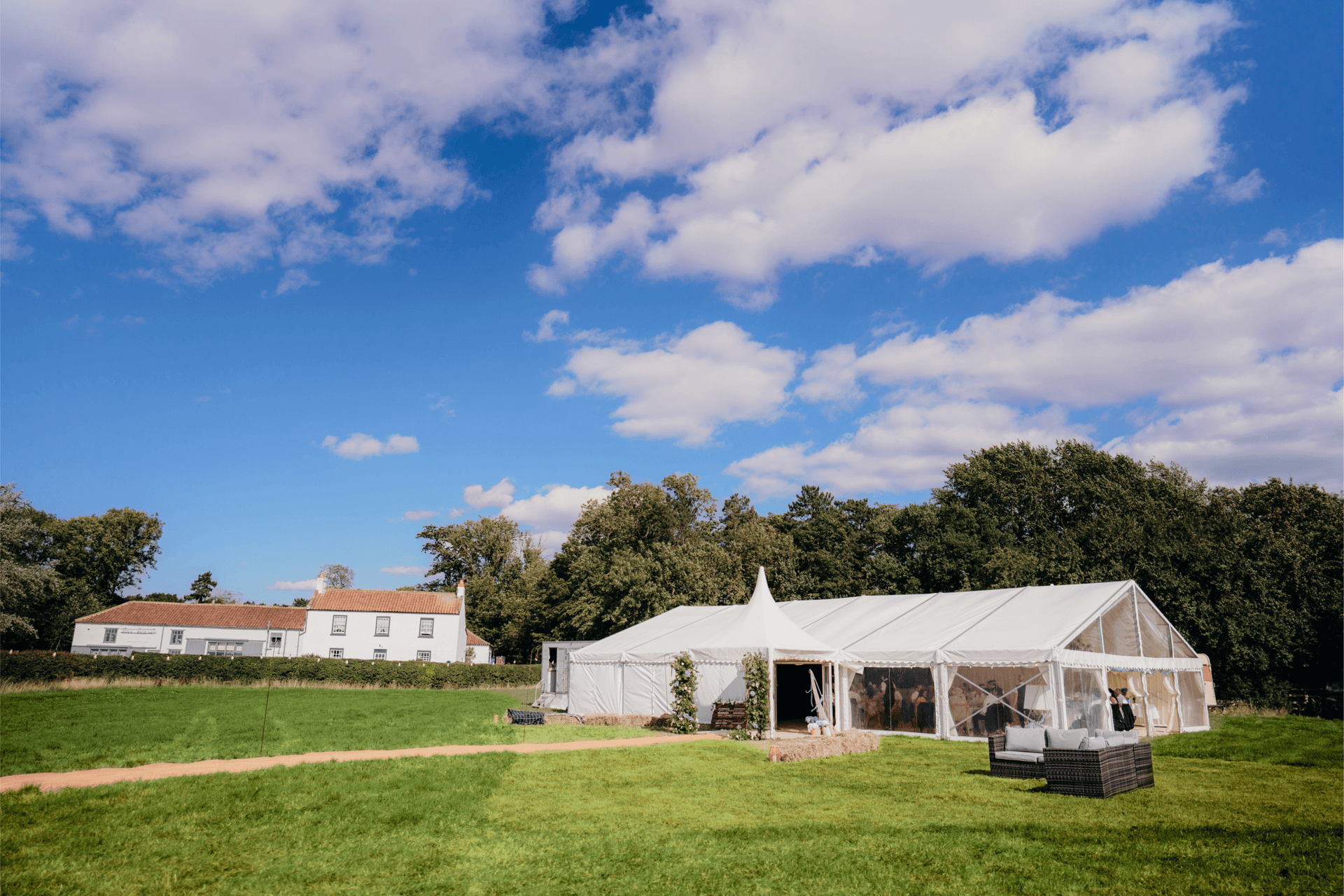 Outdoor event setup on a sunny day with large white tents on a grassy field, a white building in the background and trees beneath a blue sky with scattered clouds. Wedding Hire Near Me