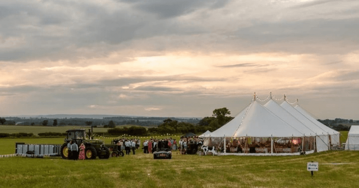 Outdoor countryside event at sunset: a large white marquee with string lights, a gathered crowd, and a tractor on the left in a grassy field.