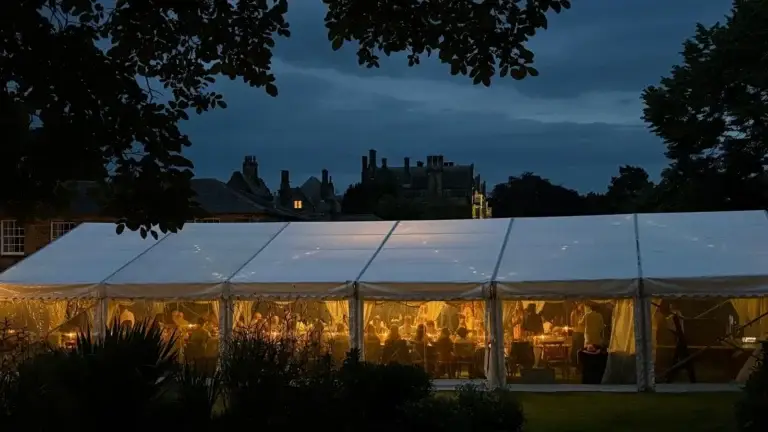 Large white event tent at dusk, warm interior lights with people dining inside, set in a garden.